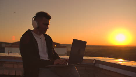 Rear-view-of-a-man-in-headphones-listening-to-music-and-working-on-the-roof-of-a-building-at-sunset-with-a-view-of-the-city-from-a-height.-Roof-of-a-skyscraper-at-sunset.